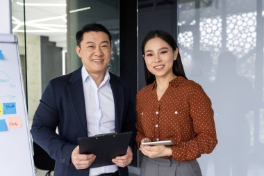 Asyalı işçiler modern ofis ortamında birlikte duruyor, takım çalışması ve profesyonelliği sergiliyorlar. Pano tutan adam, sunum için telefonu kullanan kadın, iş ortaklığı yapan adam.