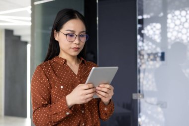 Ofiste tablet kullanan Asyalı kadın, profesyonel çevrede teknoloji kullanımını gösteriyor. Odaklanmış ifade, dijital görevler ve modern çalışma kültürüyle bağlantıyı vurguluyor.