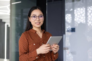 Modern ofis ortamında tablet tutan kendine güvenen Asyalı işçi. Puantiyeli bluz giymişti, arkadaş canlısı ve profesyonel tavırlar sergiliyordu, şirket ortamında çeşitli işgücünü temsil ediyordu.