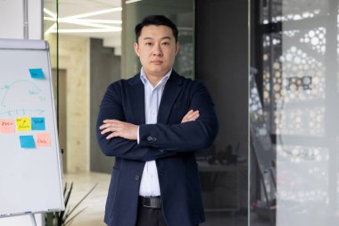 Kendine güvenen Asyalı işadamı modern ofiste kollarını kavuşturarak poz veriyor. Profesyonel kıyafet otoriteyi ve liderliği taşır. Renkli notlarla arkaplandaki beyaz tahta aktif beyin fırtınası öneriyor