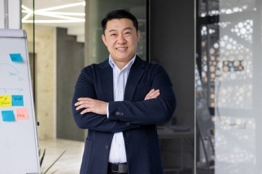 Modern ofiste kollarını kavuşturup kameraya gülümseyen kendine güvenen Asyalı bir işçi. Liderliği, profesyonelliği ve takım çalışmasını gösteriyor. Müdahale ve yakınlaşma, başarılı bir işi temsil eder.