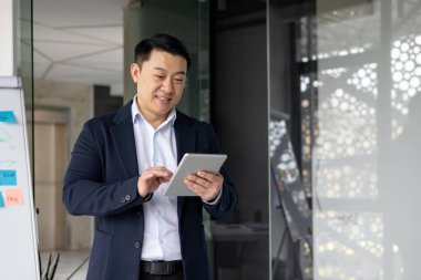 Ofis ortamında tablet kullanan Asyalı bir işçi. İş günlerinde çeşitli mesleki görevler için teknolojiyle uğraşırken gülümseyen bir adam.