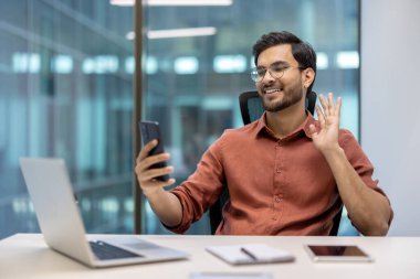 Modern ofisteki kendine güvenen genç adam video görüşmesi için akıllı telefonu kullanıyor, iş arkadaşıyla iletişim kuruyor. Teknoloji ile mutluluğu ve bağlantıyı göstermek. Uzak çalışmayı, iletişimi temsil eder