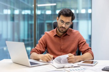 Modern ofiste oturan ciddi finansör belgeleri gözden geçirmeye odaklanıyor. İnsan kendini işine adamış ve profesyonel bir işi gösteren dizüstü bilgisayar ve telefon da dahil olmak üzere teknolojik araçlarla çevrilidir.