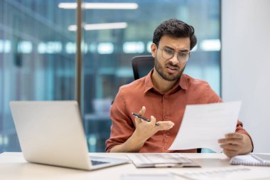 Ofis masasında dizüstü bilgisayarla oturmuş belgeyi inceleyen bir işadamı. Gözlük takıyor ve endişeli görünüyor. Eleştirel düşünme ve karar verme üzerine odaklı işyeri ayarları.