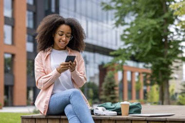 Gülümseyen Afrikalı Amerikalı kız öğrenci dışarıda otururken akıllı telefon kullanıyor. Dinlenme molası sırasında kahve ve kulaklıklar yakınlarda, mutluluk ve rahatlama hissi iletiliyor.