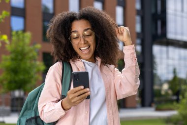 Sırt çantalı neşeli Afro-Amerikan bayan öğrenci dışarıda akıllı telefondan başarısını kutluyor. Modern akademik çevrede mutluluğu ve başarıyı gösterir.