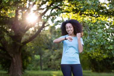 Neşeli Afrikalı Amerikalı kadın dışarıda telefon takip cihazı kullanıyor, sağlık ve sağlığa odaklanıyor. Ağaçların arasından süzülen güneş ışığı egzersiz sırasında canlandırıcı bir atmosfer yaratır..