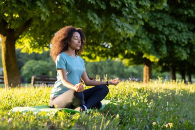 Açık havada yoga yapan Afro-Amerikalı bir kadın huzur parkında çimenlerde bacak bacak üstüne atmış oturuyor. Meditasyona dalmak, farkındalık ve rahatlamaya odaklanmak, güneşli günün sıcaklığının tadını çıkarmak..