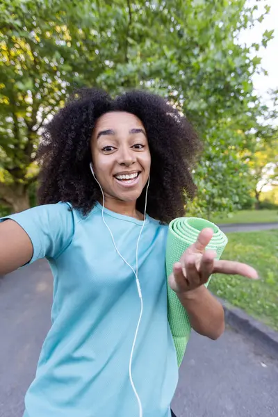 stock image Joyful African American woman enjoying outdoor fitness. Holding yoga mat, wearing headphones, engaging in video call using phone. Expresses happiness, motivation during exercise in park setting.