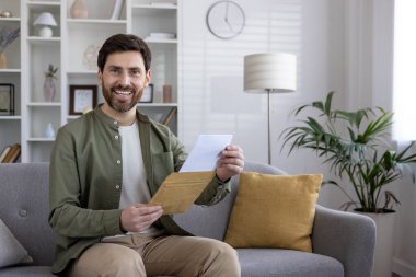 Elinde mektup zarfıyla koltukta oturan gülümseyen adam. Rahat ev ortamında olumlu haberlerin tadını çıkaran bir insan. Mutluluk, rahatlama, iletişim ve memnuniyet kavramı.