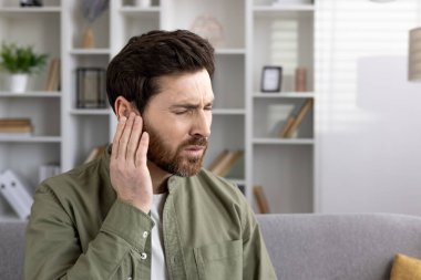 Ev ortamında koltukta oturan bir adam kulak ağrısından muzdarip. Rahatsızlık ifadesi olası kulak ağrısı ya da iltihap olduğunu gösteriyor. Gelişigüzel sağlık sorunuyla kişisel mücadele duygusunu artırır.