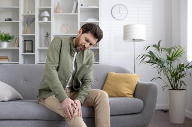 Kanepede oturan bir adam dizini rahatsız bir şekilde tutuyor. Modern oturma odası, gündelik kıyafetler. Eklem ağrısı, sağlık, yaşlanma, ev konforu kavramı.
