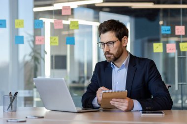 Takım elbiseli bir profesyonel laptopta özenle çalışıyor, verileri gözden geçiriyor ve ofis masasında notlar alıyor. Çevreler, cam yansıtan yaratıcı fikirler üzerine renkli yapışkan notlar içerir.