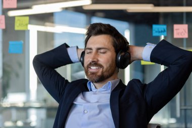 Ofisteki işadamı kulaklıklarla rahatlamanın keyfini çıkarıyor, müzik dinlerken gülümsüyor. Profesyonel kıyafet ve olumlu duygular stresten arınmış çalışma ortamını ortaya koyuyor.