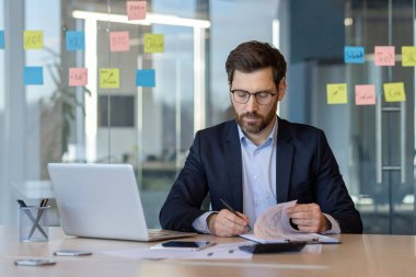 Takım elbiseli kendine güvenen bir girişimci ofis masasında bilgisayarlı ve akıllı telefonlu evrak işlerini inceliyor. Organizasyon ve planlama için renkli yapışkan notlarla çevrili, konsantre ve göreve odaklanmış..