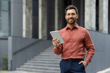 Confident businessman with tablet standing outside office building. Man wearing smart casual attire looks at camera with friendly smile. Represents technology use in professional environments clipart