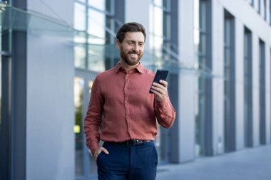 İş adamı, sıradan giyinmiş, akıllı telefona bakarken gülümsüyor. Modern ofis binasının dışında duruyor, kendine güvenini yayıyor. İletişim, teknoloji ve profesyonel yaşam tarzını yansıtır..