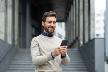 Kendine güvenen bir adam telefonu tutuyor, şirket binasının önünde dikilirken gülümsüyor. İş bağlantısını, teknolojiyi ve iletişimi temsil ediyor. Profesyonel yaşam tarzını resmetmek için ideal resim