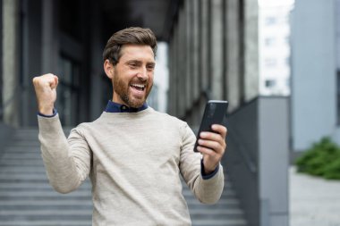 Adam akıllı telefona bakarken başarıyı kutluyor. O, mutluluk ve heyecanı yumruk pompasıyla ifade ediyor. Şehir arkaplanı onun neşeli tepkisine dinamik enerji katıyor..