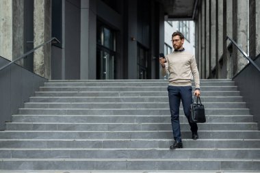 Kendine güvenen işadamı elinde telefon ve çantayla merdivenlerden iniyor. Gözlüğü, süveteri ve pantolonu var. Odaklanmış ve profesyonel görünüyor. Sahne, modern iş yaşam tarzı ve kentsel yaşamı yakalıyor