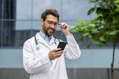 Doktor beyaz önlük ve gözlük takıyor, elinde akıllı telefonla dışarıda duruyor, telefon ekranını izlerken gülümsüyor. Sağlık uzmanlarının olumlu etkileşimini temsil ediyor