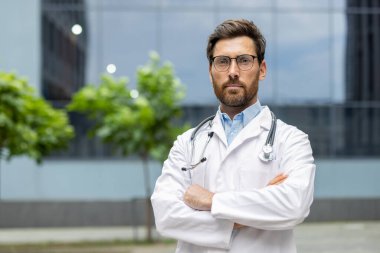 Beyaz önlüklü ve steteskoplu çapraz kollu kendine güvenen bir doktorun portresi. Profesyonelliği, tıbbi uzmanlığı ve kendini adamışlığı gösteriyor. Sağlık, hastane ve tıbbi uygulama kavramları için ideal.