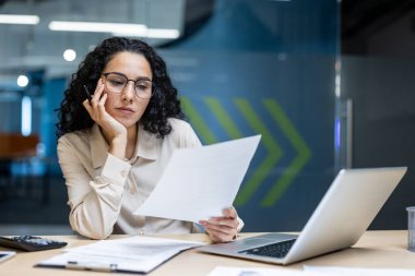 Modern ofiste masa başında oturan İspanyol iş kadını kağıtları incelerken sıkılmış görünüyor. Bir elinde belge, diğer elinde dinleniyor, gözlük takıyor. Laptop ve telefon mevcut.
