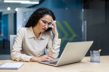 İspanyol iş kadını, modern ofis ortamında kaygı göstererek dizüstü bilgisayarda çalışmaya odaklandı. Mesleki kıyafet ve gözlükler, çalışma ortamının ortasındaki göreve bağlılığı vurguluyor