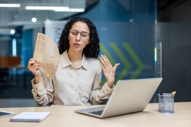 İspanyol iş kadını ofis masasında dizüstü bilgisayar kullanarak oturuyor ve vantilatörle serinliyor. Sıcaktan rahatsız olduğunu belirterek havalandırma eksikliği olduğunu öne sürüyor..
