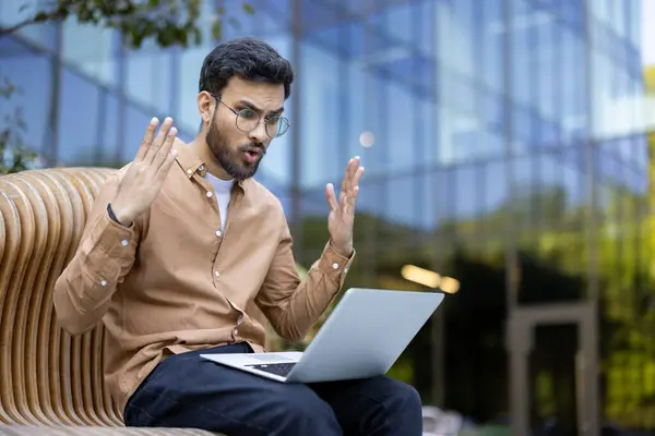 stock image Young man sitting outdoors, displaying frustration with laptop. Occurs in urban environment showcasing unexpected technical problem. Concept of technology mishap, communication, and workplace stress