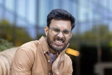 Gözlük takan genç adam diş ağrısından dolayı acı çekiyor, yanağını rahatsızlık içinde tutuyor. Modern ofis binasının dışında yakalanmış, diş sorunu ve iş günlerinde yaşanan rahatsızlığı resmediyor..