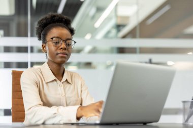 Modern ofiste dizüstü bilgisayar kullanarak gözlük takan Afro-Amerikalı bir iş kadını. İşine odaklanmış ve meşgul görünüyor, profesyonelliğini ve rolüne bağlılığını gösteriyor. Temiz ortam