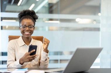 Genç afro-amerikan kadın gülümseyen ve başarıyı kutlayan bir şekilde ofis masasında dizüstü bilgisayarla telefon ediyor. İş ortamında başarı, kutlama ve teknoloji kavramı.