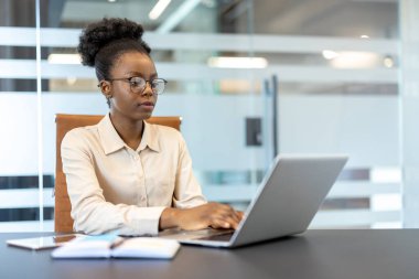 Genç Afrikalı Amerikalı iş kadını, modern ofis ortamında iş yerinde dizüstü bilgisayarla çalışırken odaklandı. Verimlilik ve profesyonelliği gösteren ciddi ve konsantre ifade.