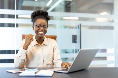 Genç Afrikalı Amerikalı iş kadını dizüstü bilgisayar kullanıyor, gülümsüyor ve başarısını gösteriyor. Modern teknoloji ve kararlı ifade ile profesyonel çalışma ortamı