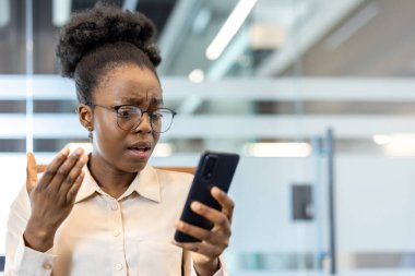 Ofisteki Afrikalı Amerikalı genç iş kadını telefona şaşkın şaşkın bakıyor. Gözlük ve beyaz gömlek takmış, telefonunda gördüğü bilgilerle kafası karışmış ya da şok olmuş gibi görünüyor..