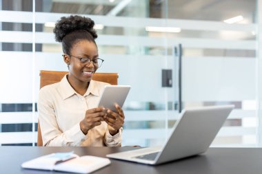 Modern ofiste tablet kullanırken gülümseyen genç Afrikalı Amerikalı iş kadını. Masasında dizüstü bilgisayarı ve defteri olan dijital bir proje üzerinde çalışan profesyonel bir çalışan.