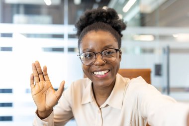 Ofisteyken kameraya el sallayan gözlüklü gülümseyen kadın. Telefonla kameraya bakarken mutlu profesyonel selamlaşmalar. Sanal toplantılar ve uzaktan iletişim kavramı.
