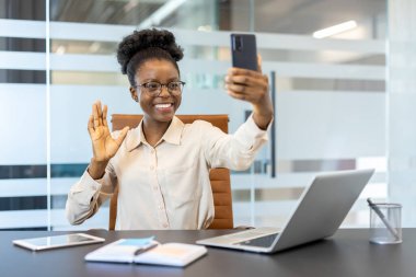 Genç Afrikalı Amerikalı iş kadını video görüşmesi için telefonu kullanıyor, gülümsüyor ve modern ofisin iş yerinde elinde laptop ve belgelerle el sallıyor.