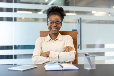 Kendinden emin genç Afrikalı Amerikalı iş kadını modern ofis masasında oturmuş kameraya gülümsüyordu. Ofis malzemeleri ve telefon masadaki verimliliği gösteren çalışma ortamında.
