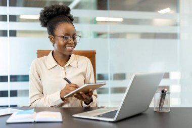 Ofis masasında dizüstü bilgisayarı ve not defteriyle çalışan genç Afrikalı Amerikalı iş kadını görevlere odaklanıyor. Profesyonel kıyafet, gülümseme ve görünüşünü güzelleştiren gözlük. Verimlilik kavramı