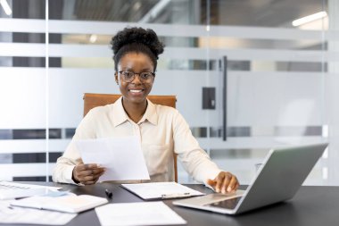 Afrikalı Amerikalı iş kadını modern ofis ortamında masabaşında güvenle çalışıyor. Belgeleri elinde tutuyor ve dizüstü bilgisayar kullanıyor. Profesyonelliğini gösteriyor ve odaklanıyor..