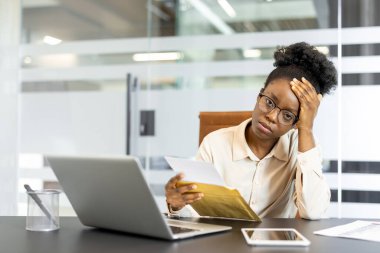 Afro-Amerikalı iş kadını, ofis masasında kötü haberler aldıktan sonra stresli hissediyor. Laptop ve telefonla oturuyor, zor durumu düşünüyor..