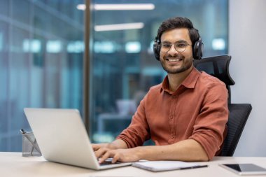 Modern ofis ortamında dizüstü bilgisayar kullanan gözlüklü ve kulaklıklı genç bir profesyonel. Dijital çalışmayla meşgul olan kişi kendinden emin bir şekilde gülümsüyor. Ofis ortamı yaratıcılığı ve odaklanmayı teşvik eder