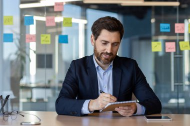 Modern ofisteki iş adamı odaklı ifadeyle notlar yazıyor. Cam duvardaki görünür yapışkan notlar üretkenliği ve yaratıcılığı vurguluyor. Yakın telefon iş iletişimini öneriyor.