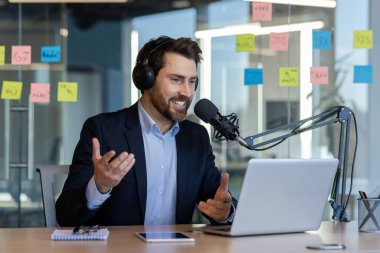 Podcast 'i ofiste mikrofon, kulaklık, dizüstü bilgisayar ile kaydeden adam, coşku ve profesyonelliği ifade ediyor.