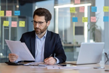 Masada çalışan, gözlük takan, belgeleri analiz eden profesyonel bir iş adamı. Modern ofis ortamında dizüstü bilgisayar ve akıllı telefon, cam duvarda yapışkan notlar..