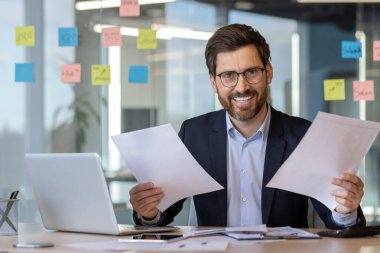 Ofis masasında bilgisayarlı ve telefonlu belgeleri incelerken gülümseyen, kendine güvenen işadamı..