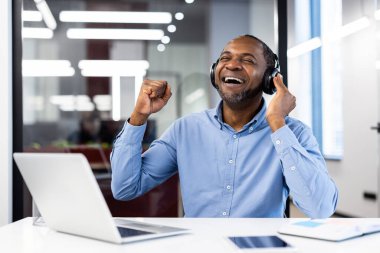 Günlük ofis ortamında gülümseyen adam kulaklık takıyor, müzikten zevk alıyor ve başarıyı kutluyor. Masadaki dizüstü bilgisayar ve telefon teknoloji odaklı çalışma ortamını vurguluyor.